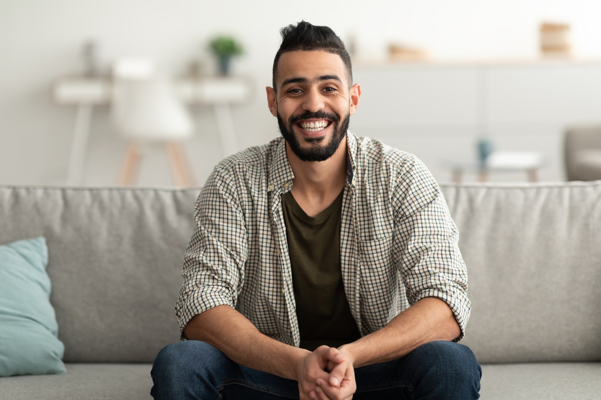 Portrait of handsome young Arab man smiling and looking at camera, sitting on sofa at home